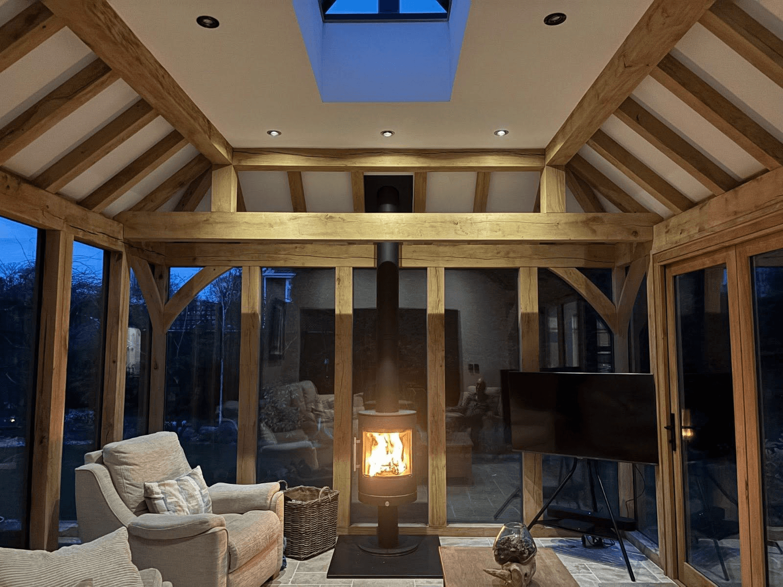 Interior of an oak-framed garden room at dusk — exposed green oak frame and vaulted roof, glazed walls and a wood-burning stove