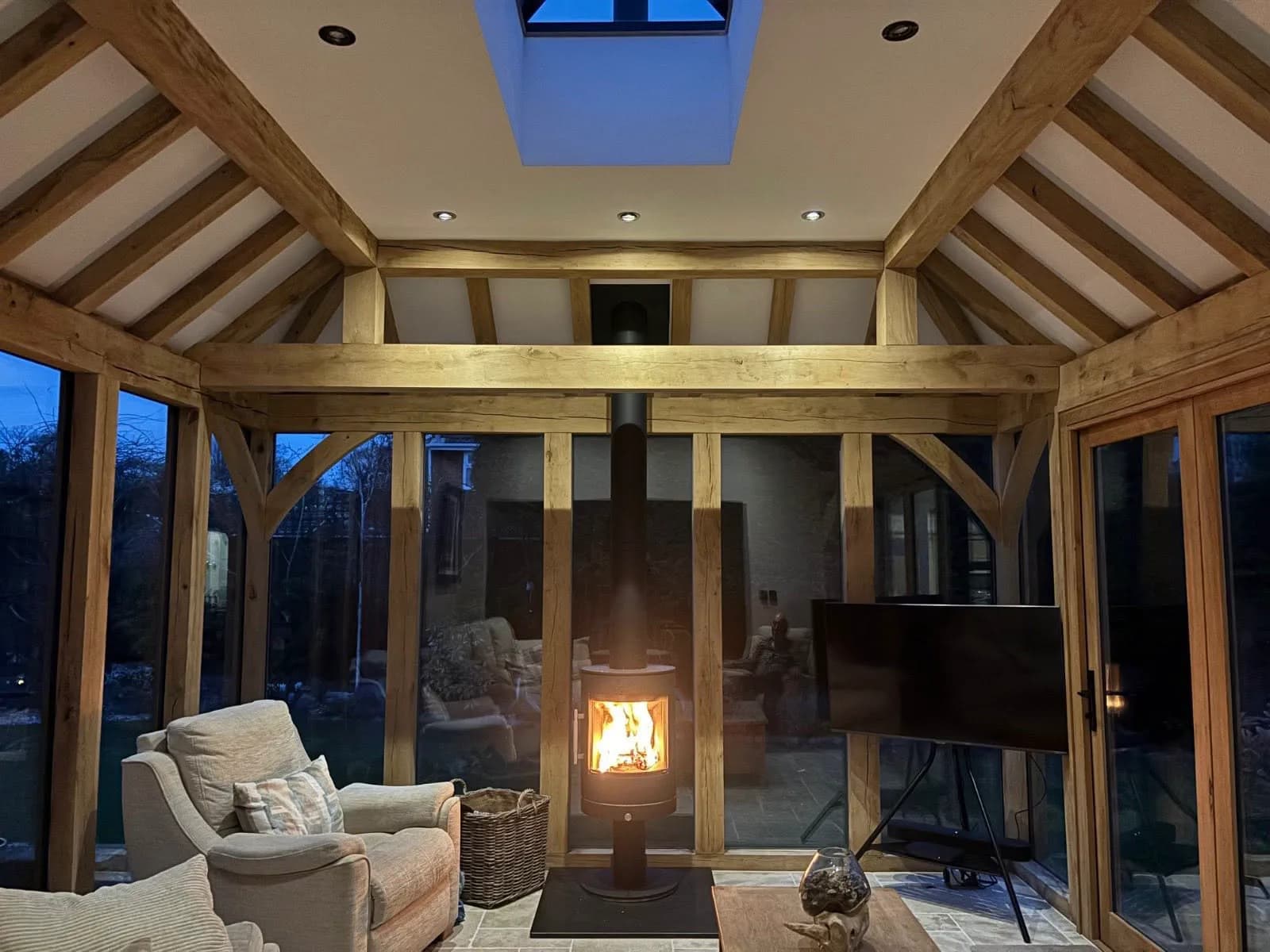 Interior of an oak-framed garden room at dusk — exposed green oak frame and vaulted roof, glazed walls and a wood-burning stove