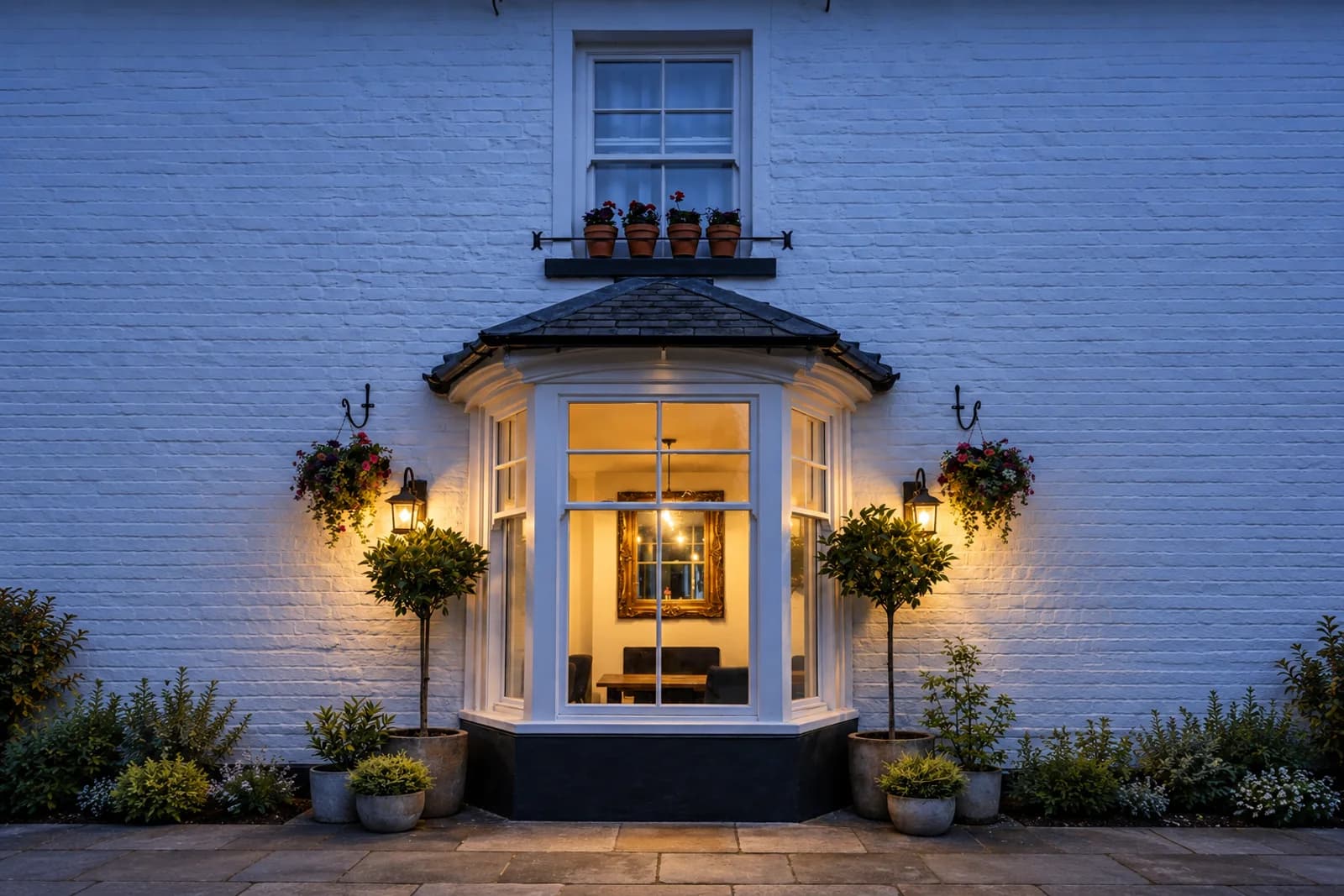 Canted bay window lit from within at dusk beneath a slate bay roof, between wall lanterns on a painted brick elevation, with clipped topiary either side and a sash window above