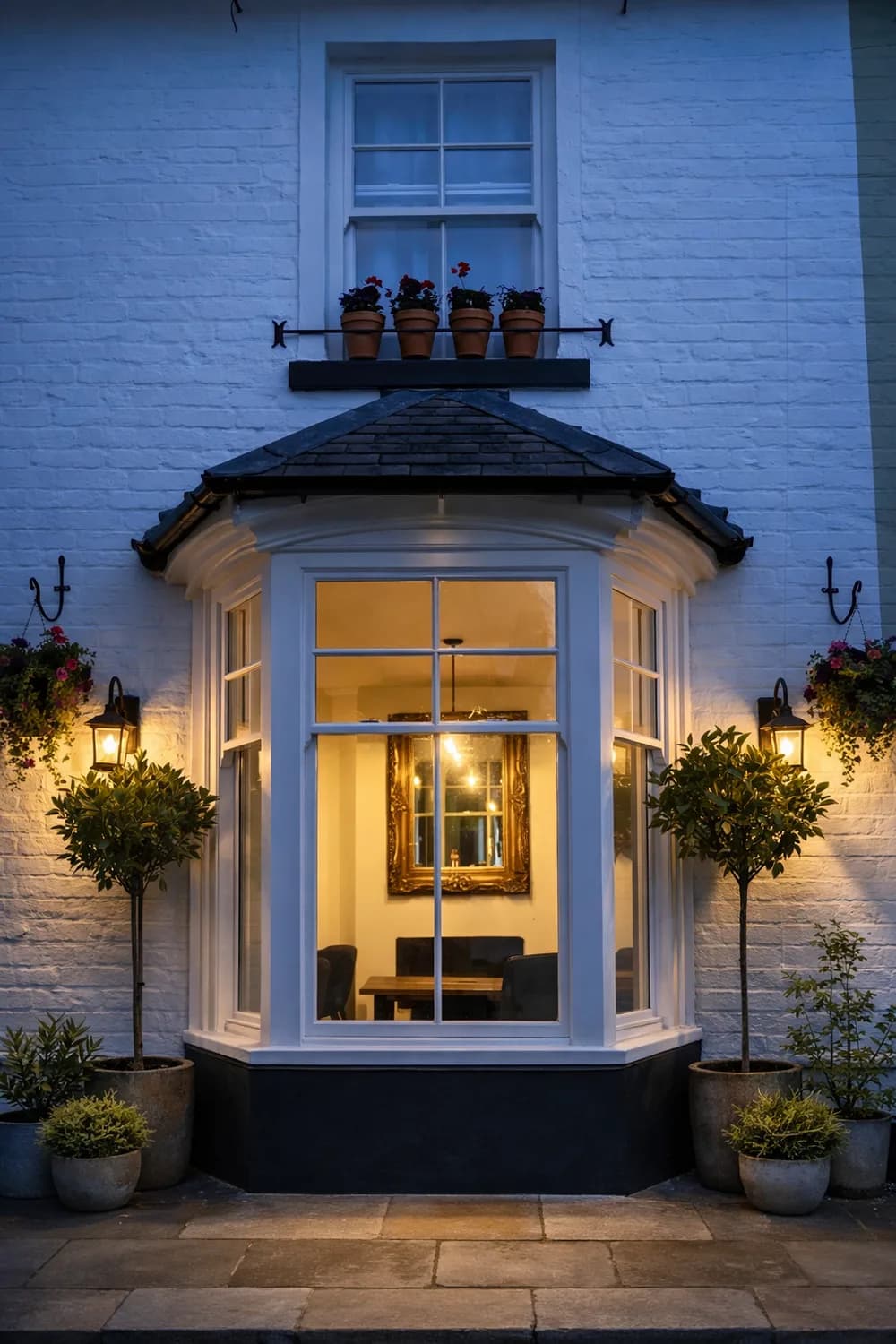 Canted bay window lit from within at dusk, under a slate bay roof on a painted brick elevation, with bay trees either side