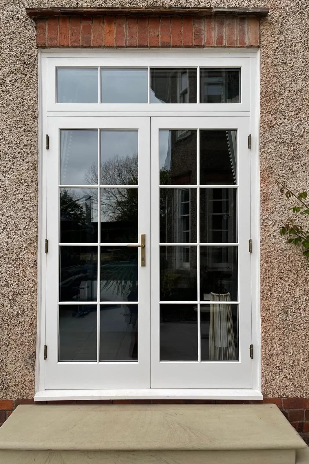 White-painted Accoya French doors with a transom light above, multi-pane glazing and bronze ironmongery, in a roughcast elevation with a brick lintel