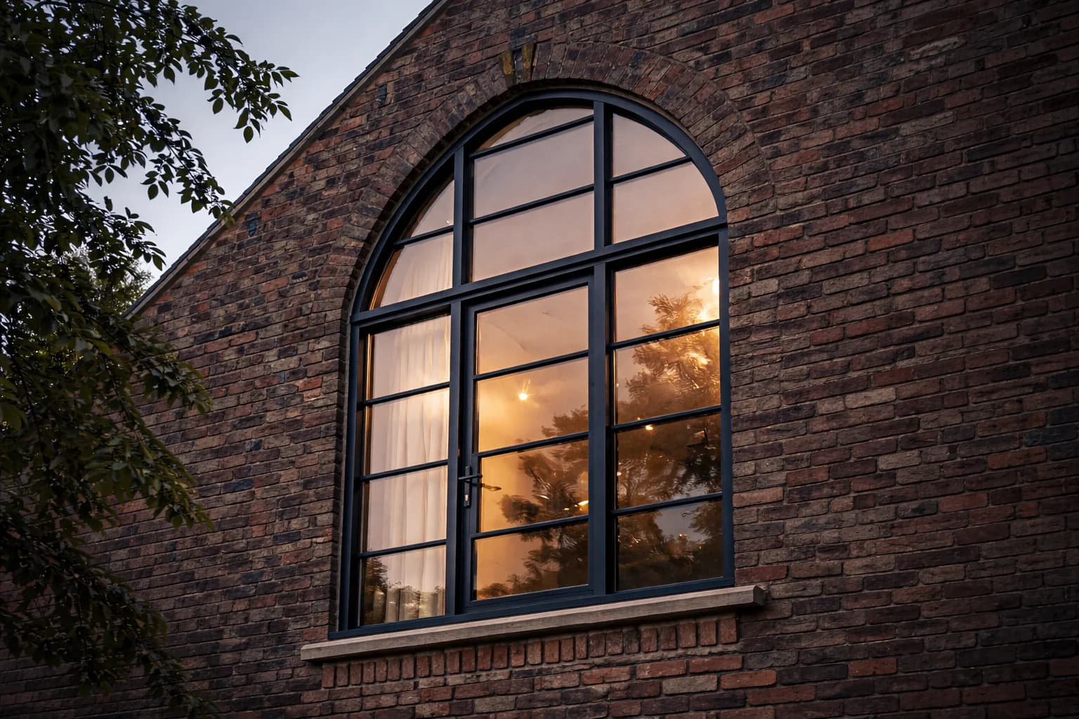 Arched steel-look Accoya window in the gable of a converted brick barn at dusk, its head following the brick arch, the room lit behind it
